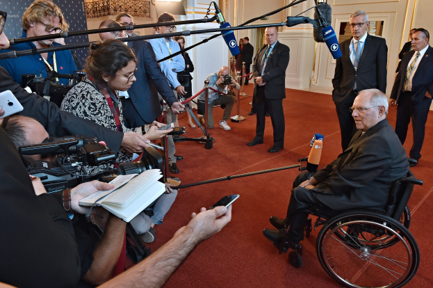 Ein Mann im Rollstuhl ist von Reportern umgeben, die Kameras, Handys und Mikrofone mit Ständern halten, sowie ein Banner, Scheinwerfer und eine Wand im Hintergrund.