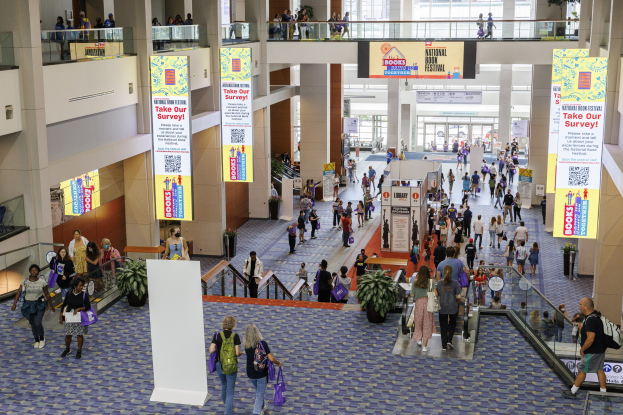 Eine große Menschenmenge, die durch ein großes Gebäude mit Bannern, Topfpflanzen, Rolltreppen, Geländern, Säulen, Texttafeln, Glastüren und anderen Gegenständen geht, wobei einige Taschen tragen, wahrscheinlich an der Nationalen Buchmesse teilnehmen.