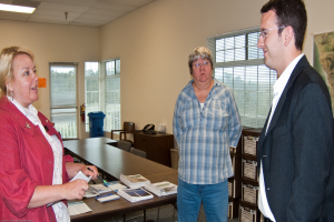 Eine Frau in einer roten Jacke hält einen Stift und Papier in der Hand und steht links neben einer Frau in einem blauen karierten Hemd und einem Mann in einem weißen Hemd und einem schwarzen Anzug mit Brille, alle in der Nähe eines Tisches mit Papieren, eines Schreibtisches mit einem Telefon, eines Stuhls, eines roten Feuerlöschers und einer Kiste in der Ecke.