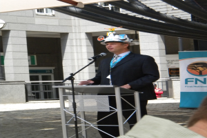 Ein Mann in Anzug und Hut spricht in ein Mikrofon an einem Pult, mit einer Person rechts unten, einem Banner mit Text rechts und einem Gebäude mit Gelönder im Hintergrund.