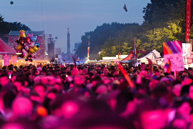 Eine große Menschenmenge auf einem nächtlichen Musikfestival mit farbiger Beleuchtung, umgeben von Zelten, Fahnen und Pfählen, mit Bäumen, einem Turm und einem klaren Himmel im Hintergrund.