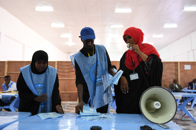 Menschen versammeln sich an einer Wahllokal um einen Tisch mit einem Megafon und Papieren, mit anderen im Hintergrund auf Stühlen und Stehenden unter beleuchteter Decke.