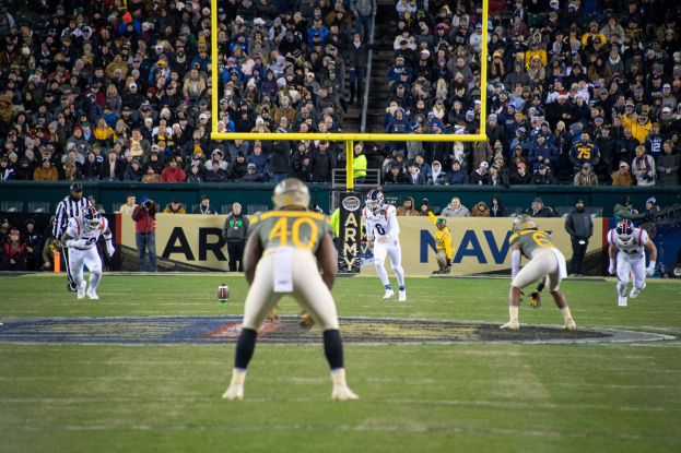 Fussballspiel in einem Stadion mit Spielern in Sportkleidung und Helmen im Vordergrund und Zuschauern im Hintergrund, mit sichtbaren Stadionwerbung und Pfählen.