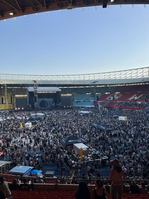 Große Menschenmenge auf Tribünen und im Stehen in einem Stadion mit Zelten, Geländern und Überdachungsleuchten vor einem Himmel als Hintergrund.