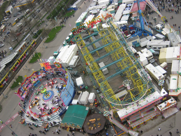 Luftaufnahme eines Vergnügungsparks mit einem zentralen Riesenrad, umgeben von Fahrgeschäften, Gebäuden, Fahrzeugen, Menschen, Straßen, Bäumen und anderen Parkmerkmalen.