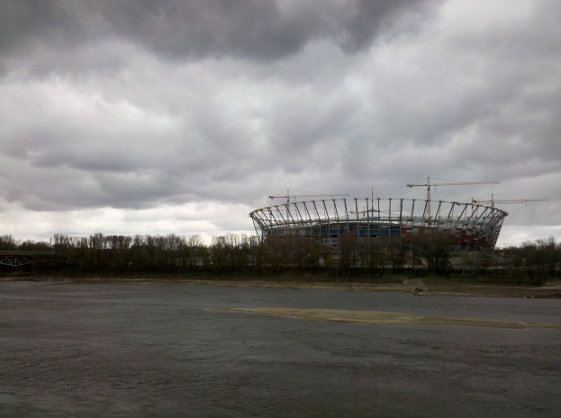 Das Olympiastadion in Kiew, Ukraine, ein großer Stadion umgeben von Bäumen und einer Brücke, mit einem bewölkten Himmel im Hintergrund und sichtbarem Boden unten.