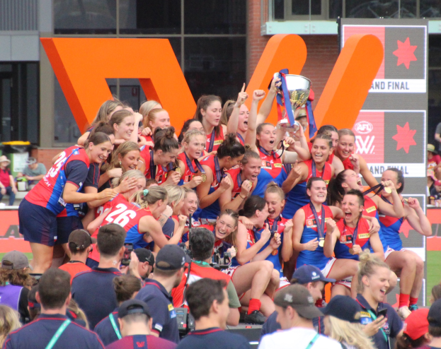 Eine Gruppe von Frauen-AFL-Spielerinnen feiert mit einem Pokal vor einem Publikum, trägt Sportuniformen und hält den Pokal in den Händen, mit einem Glasfenster-Gebäude und einer Texttafel im Hintergrund.