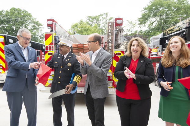 Eine Gruppe von Menschen, die bei einer Einweihungszeremonie der Alexandria Fire Department klatschen und lächeln, während zwei Personen eine Schere und ein rotes Tuch halten.