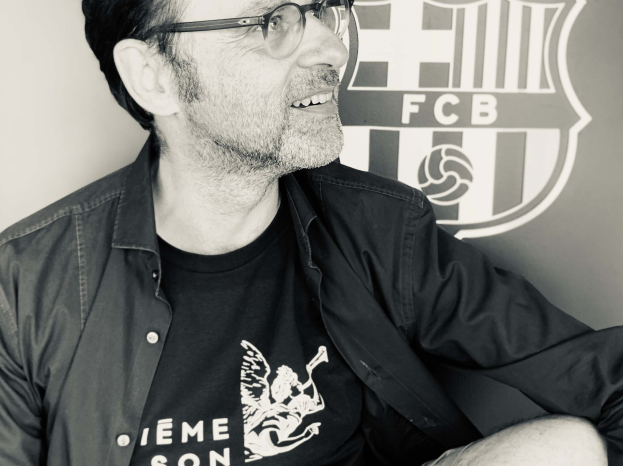 Schwarzes und weißes Foto eines grinsenden Mannes in Brille und schwarzem Hemd vor dem FC Barcelona-Logo.