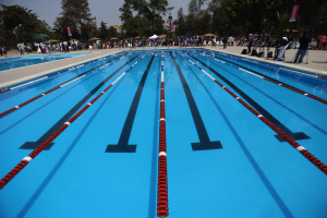 Großer Schwimmbad mit Bahnentrennern, umgeben von Menschen mit Taschen und Gegenständen, mit Zelten, Transparenten, Stangen, einer Straßenlaterne, einem Schild und einem bewölkten Himmel im Hintergrund.