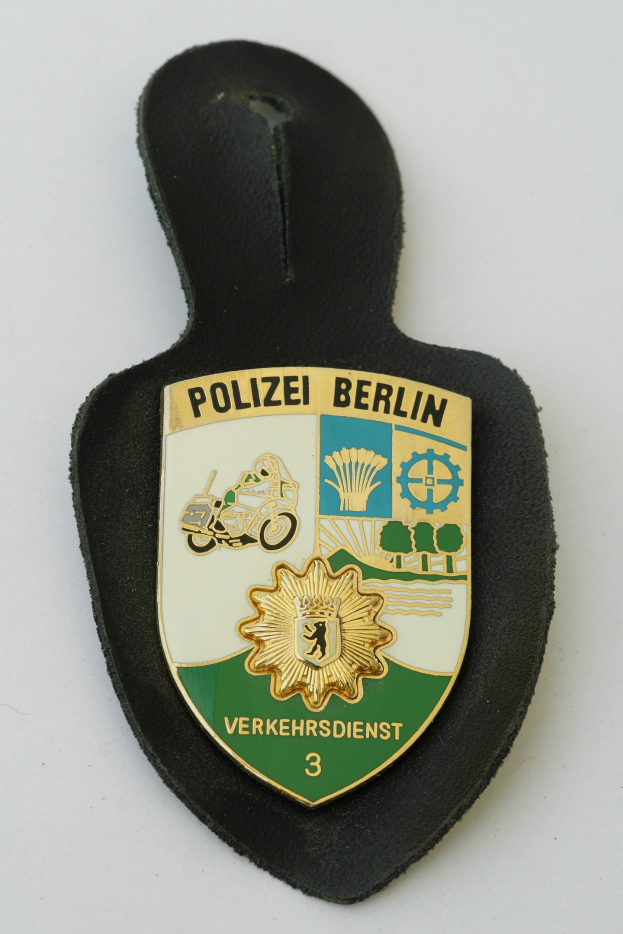 Ein schildförmiges Polizeibadge mit 'Polizei Berlin' in schwarzer fettgedruckter Schrift auf einem blauen Hintergrund mit einem weißen Stern und Rand, das auf einer Oberfläche ruht.