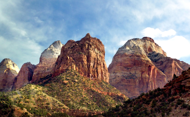 Panoramablick auf den Zion-Nationalpark in Utah mit majestätischen Bergen, grünen Bäumen, felsigem Gelände und einem Himmel mit weißen, flauschigen Wolken.
