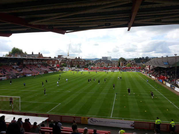 Ein Fußballspiel im St. Mary's Stadium mit Zuschauern auf den Tribünen und in der Nähe des Torpfostens, umgeben von Gebäuden, Bäumen und einem bewölkten Himmel.