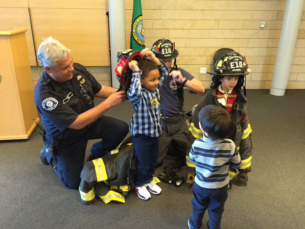 Eine Gruppe von Kindern in Feuerwehranzügen und ein Feuerwehrmann in Uniform stehen um ihn herum, während er einen Helm hält, mit einer Tasche auf dem Boden in einer Feuerwache.