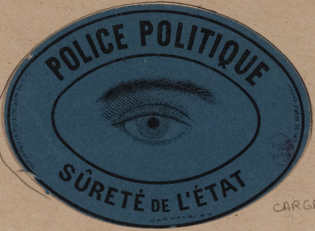 Brauner Karton mit einem blauen Aufkleber, auf dem der Text 'Polizei Politique Sûreté de l'État' und ein zentrales Auge-Symbol steht.