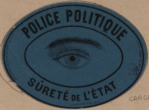 Brauner Karton mit einem blauen Aufkleber, auf dem der Text 'Polizei Politique Sûreté de l'État' und ein zentrales Auge-Symbol steht.