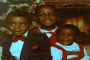 Drei Kinder lachen vor einem Weihnachtsbaum mit einem Kamin aus Holz auf der rechten Seite.