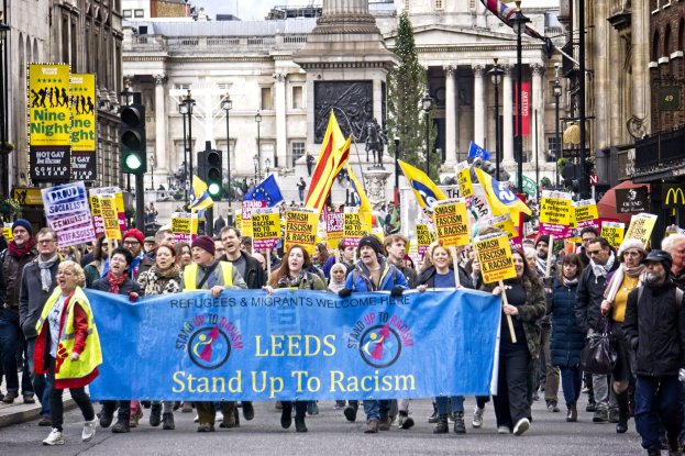 Eine Gruppe von Menschen marschiert mit einer 'Steh Auf gegen Rassismus'-Plakette die Straße entlang, mit Gebäuden, Bäumen, Laternen, Ampeln und einer Statue im Hintergrund bei bewölktem Himmel.