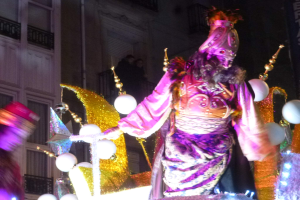 Ein Mann in einem Kostüm steht auf einem geschmückten Festwagen bei einem Umzug, im Hintergrund ist ein Stadtgebäude mit Fenstern und Geländern zu sehen.