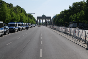 Eine Reihe von Polizeiwagen, die auf einer Straße vor dem Brandenburger Tor in Berlin, Deutschland, geparkt sind, mit Menschen auf Fahrrädern und auf der Straße stehend, Barrieren, Bäumen und einem Bogen mit Statuen im Hintergrund.