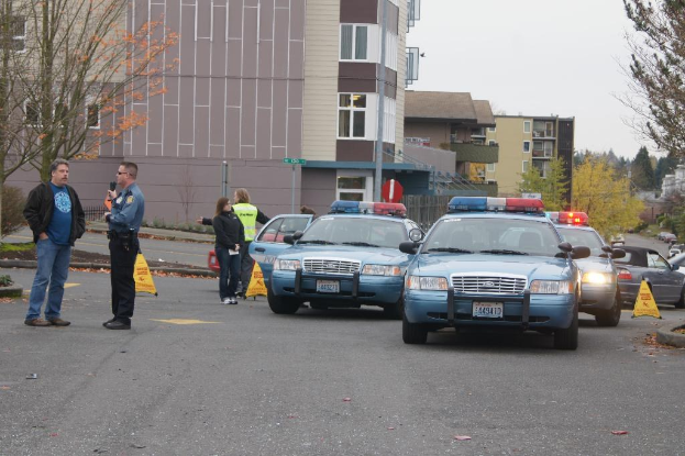 Vier Autos auf einer Straße, vier Personen in der Nähe, Gebäude mit Fenstern im Hintergrund, Bäume und ein Warndreieck.