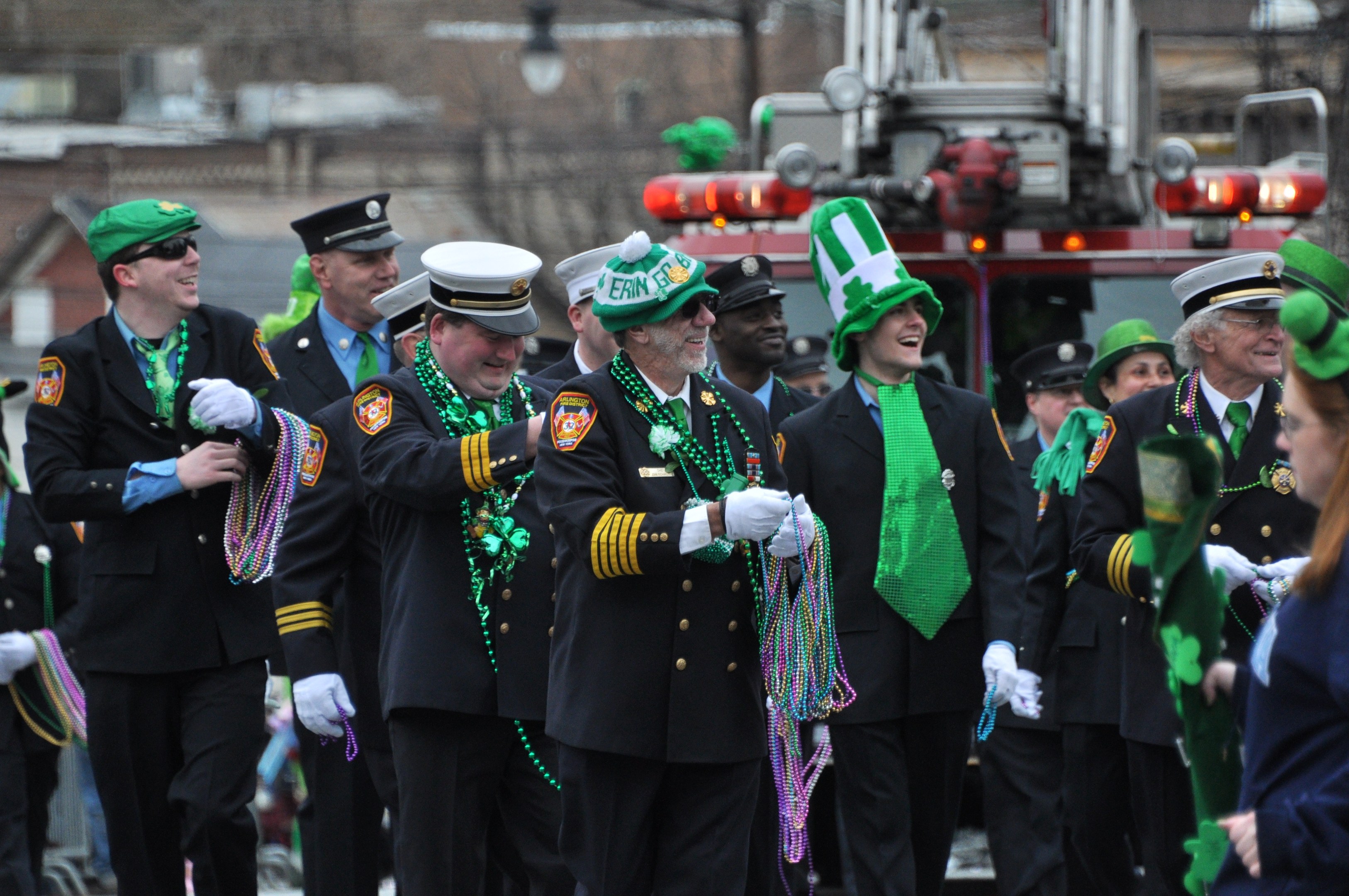 Eine Gruppe von Männern in Uniform marschiert in einem St. Patrick's Day Umzug, hält Perlen, mit einem Feuerwehrauto und Gebäuden im Hintergrund.