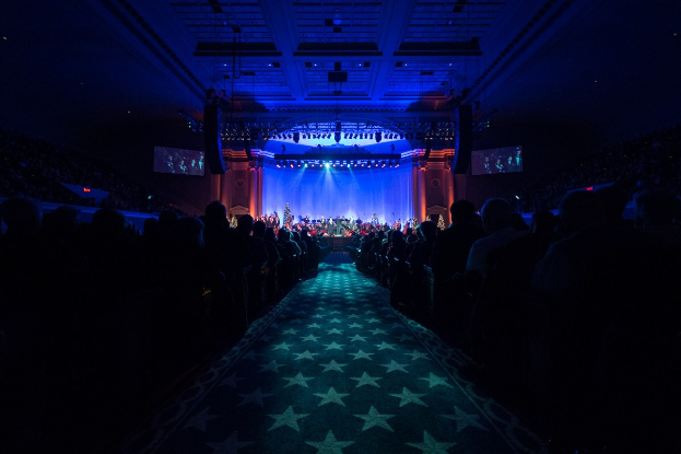 Ein großer, dämmrig beleuchteter Zuschauerraum mit Sitzplätzen, eine Bühne mit Musikern und Überkopfbeleuchtung und -fernseher, was auf einen Konzerthallensaal während der Nationalkonvention 2019 hinweist.