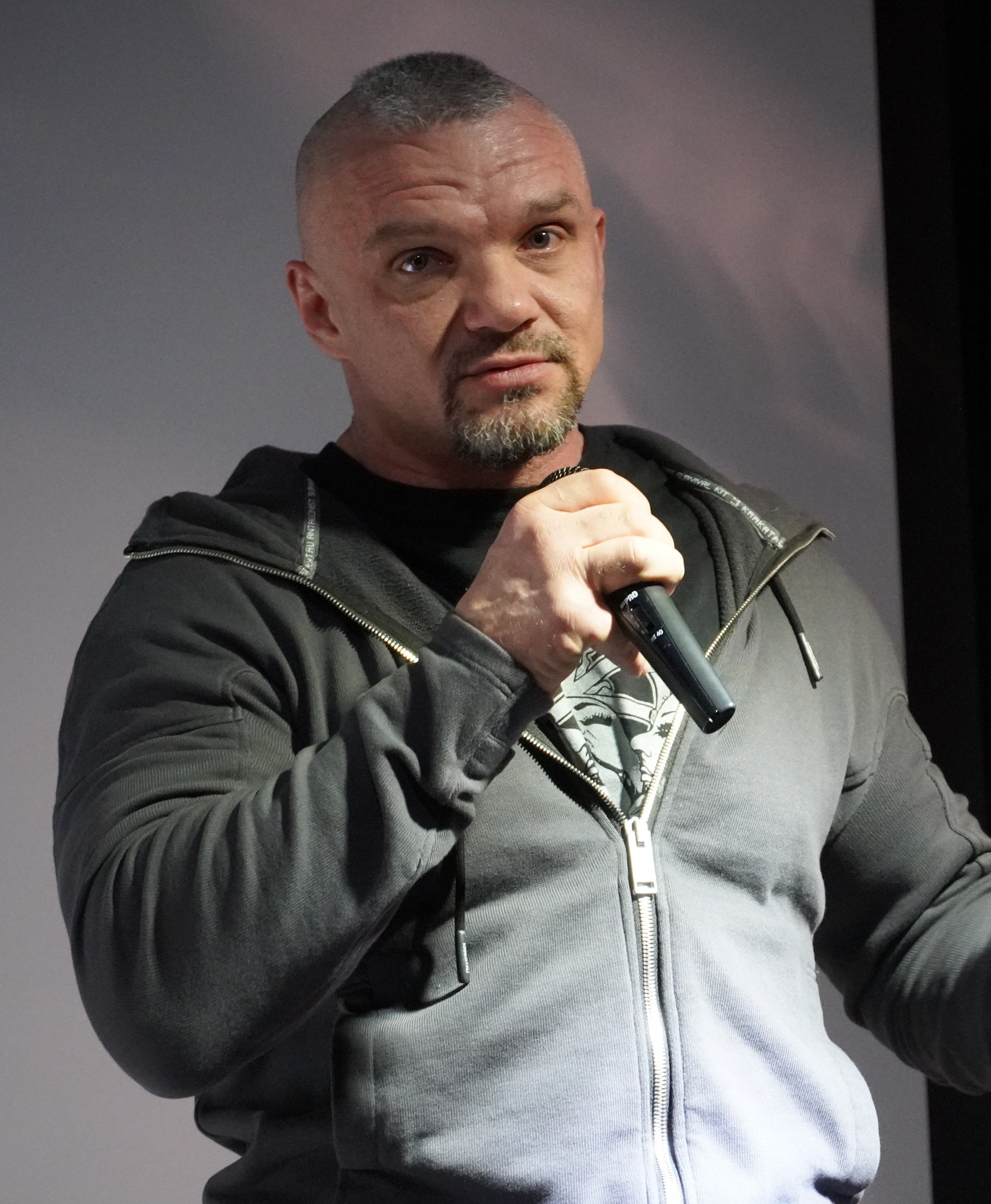 Ein Mann in schwarzer Kapuzenjacke und schwarzem Anzug mit weißem Hemd hält ein Mikrofon und steht selbstbewusst vor einer Wand, wobei er wie ein Komiker wirkt.