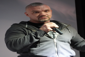 Ein Mann in schwarzer Kapuzenjacke und schwarzem Anzug mit weißem Hemd hält ein Mikrofon und steht selbstbewusst vor einer Wand, wobei er wie ein Komiker wirkt.