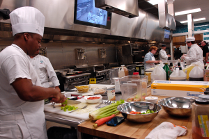 Eine Gruppe von Köchen in einer Küche, wobei einer im Vordergrund einen weißen Kittel und eine Haube trägt, ein Messer hält und von Utensilien, Schüsseln, Gemüse, einem Handy, Flaschen und anderen Gegenständen umgeben ist, während im Hintergrund ein Fernseher und Deckenleuchten zu sehen sind.