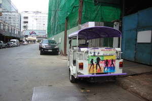 Gebäude mit parkenden Autos, eine Werbetafel und ein bewölkter Himmel, mit einem Auto-Rikscha am Rand.