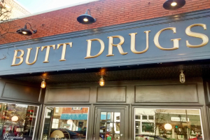 Außensicht eines Gebäudes mit einer beleuchteten Schilder 'butt drugs', das von Glaswänden und -türen umgeben ist, mit Fahrzeugen, Gebäuden, Bäumen und Himmel im Hintergrund.