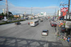 Eine belebte Stadtstraße mit verschiedenen Fahrzeugen, Strommasten, Gebäuden, Bäumen und einem Turm unter einem bewölkten Himmel.