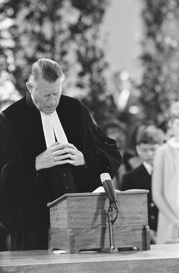 Ein Richter in einer schwarzen und weißen Robe spricht an einem Podium mit einem Mikrofon, umgeben von ein paar Menschen und Bäumen im Hintergrund.