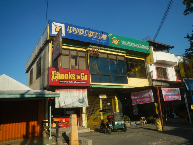 Außenansicht eines Gebäudes mit einer 'Advance Credit Corp'-Aufschrift, umgeben von Fahrzeugen, Strommasten, Bäumen und Werbetafeln unter einem klaren blauen Himmel.