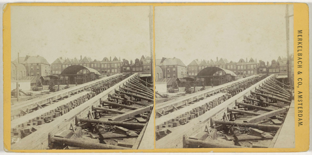 Schwarz-weißes Foto einer Zugstrecke mit Gebäuden im Hintergrund, Text "Merkelbach & Co, Co. Amsterdam" auf der rechten Seite und verstreute hölzerne Gegenstände im Vordergrund.