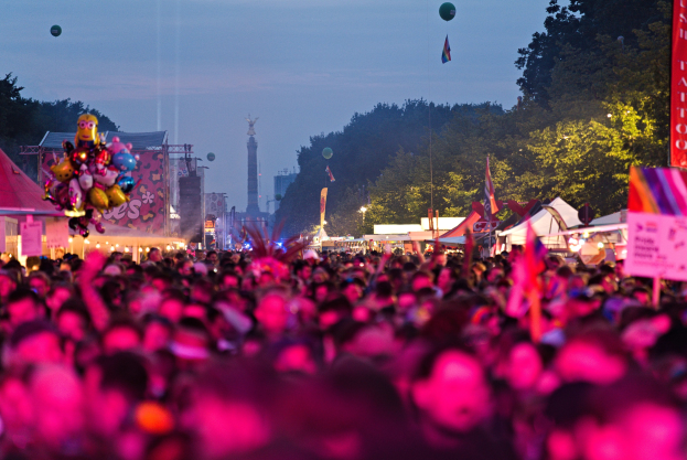 Eine große Menschenmenge auf einem nächtlichen Musikfestival mit bunten Lichtern und Ballons, umgeben von Zelten, Bannern, Fahnen, Bäumen, einem Turm und einem sternenklaren Himmel.