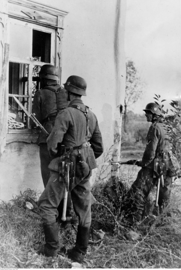 Schwarzes und weißes Foto von deutschen Soldaten in Helmen mit Gewehren, die vor einem Gebäude mit einem Fenster stehen, Bäume sind im Hintergrund zu sehen.