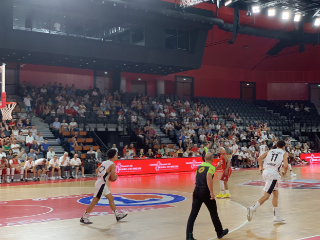 Eine Gruppe von Menschen, die Basketball in einer Turnhalle mit einem Netz auf der linken Seite spielen, Zuschauer auf Stühlen und Deckenbeleuchtung.
