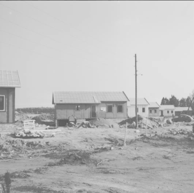 Schwarz-weiß-Foto einer Schotterstraße mit Häusern, Bäumen und Strommasten im Vordergrund und Himmel im Hintergrund, beschriftet als Baustelle für ein neues Haus.