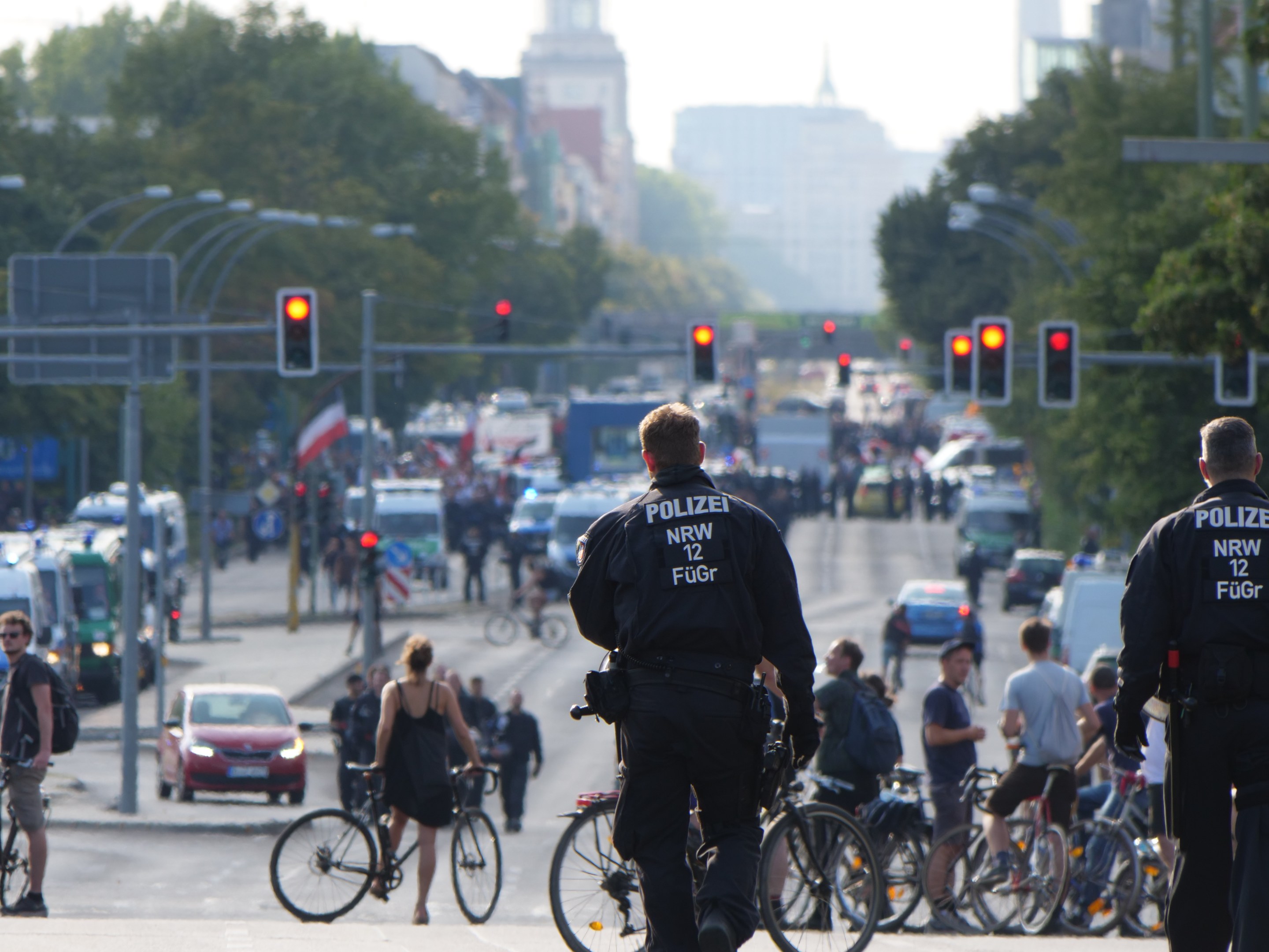 Zwei Polizeibeamte stehen an der Straße neben einer Gruppe von Radfahrern, mit Fahrzeugen, Verkehrszeichen, Bäumen, Gebäuden und einem klaren blauen Himmel im Hintergrund.