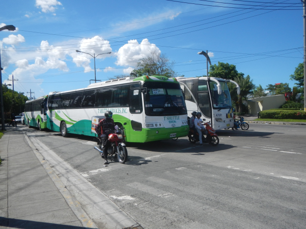 Ein grüner und weißer Shuttlebus parkt am Straßenrand mit Motorrädern, die davor vorbeifahren, umgeben von einem grasbewachsenen Gehweg und Laternenpfählen, mit Bäumen, Gebäuden und einem klaren blauen Himmel im Hintergrund.
