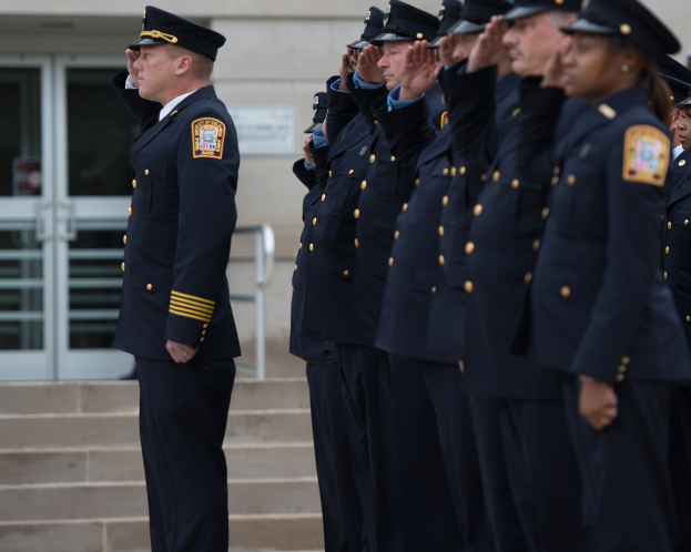 Gruppe von Polizisten in Uniform und Mützen, die in Formation vor einem Gebäude mit Glastüren und Treppen salutieren.