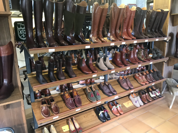 Eine Ladeneinrichtung mit verschiedenen Reitstiefeln mit Preisschildern, mit einem Stuhl auf der rechten Seite und zusätzlichen Artikeln auf den Regalen im Hintergrund.