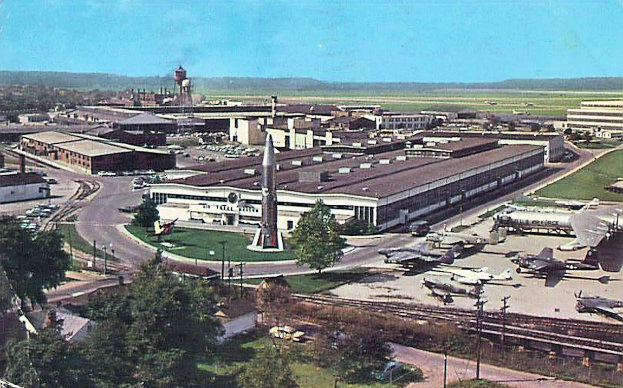 Altes Schwarz-Weiß-Foto eines Flughafens mit geparkten Flugzeugen, Gebäuden, Bäumen, Straßen, Fahrzeugen, Strommasten, Gras, Bahnschienen, Türmen, fernen Hügeln und einem klaren blauen Himmel; Text erscheint unten.