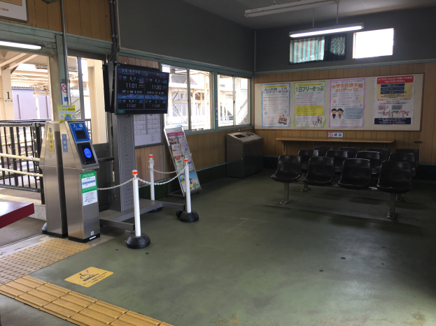 Ein belebter Bahnhofseingang mit Reihen von Stühlen, einer Service-Theke, Ticketautomaten, Kettengliedern, Informationsanzeigen, Wandplakaten, Deckenbeleuchtung und Geländern.
