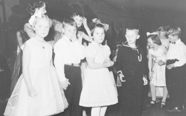 Schwarzes und weißes Bild einer Gruppe von Kindern, die auf einer Tanzfläche stehen und fröhlich und feierlich aussehen, mit einer Wand im Hintergrund.