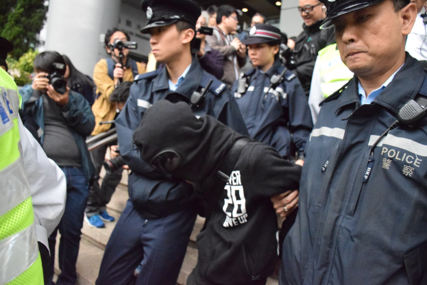 Polizeibeamte vor einer Menge mit Kameras neben einem Gebäude mit Säulen und Deckenbeleuchtung, Bäume links sichtbar.