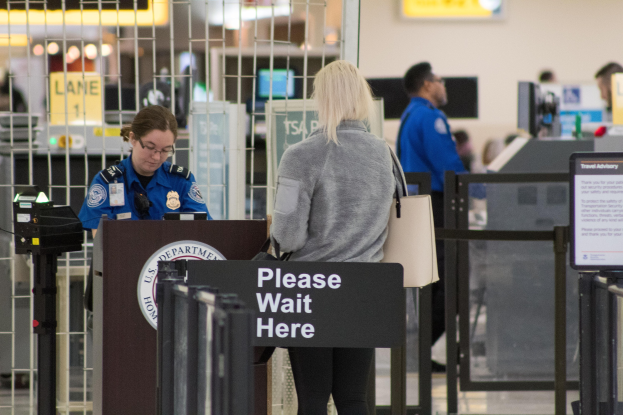 Eine Frau in einer grauen Jacke und schwarzen Hosen steht an einem Flughafen-Metallsensor, trägt eine Handtasche und hat einen Polizeibeamten in einer blauen Uniform links von sich und eine schwarze Tafel hinter sich.