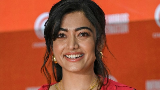 Eine Frau in einem roten Kleid mit einem eleganten Dutt und dezentem Make-up lächelt in die Kamera, trägt schwarze High Heels und hat einen hellgelben Banner mit der Aufschrift 'Sonam Kapoors beste Schönheits Looks vom Cannes Film Festival' im Hintergrund.